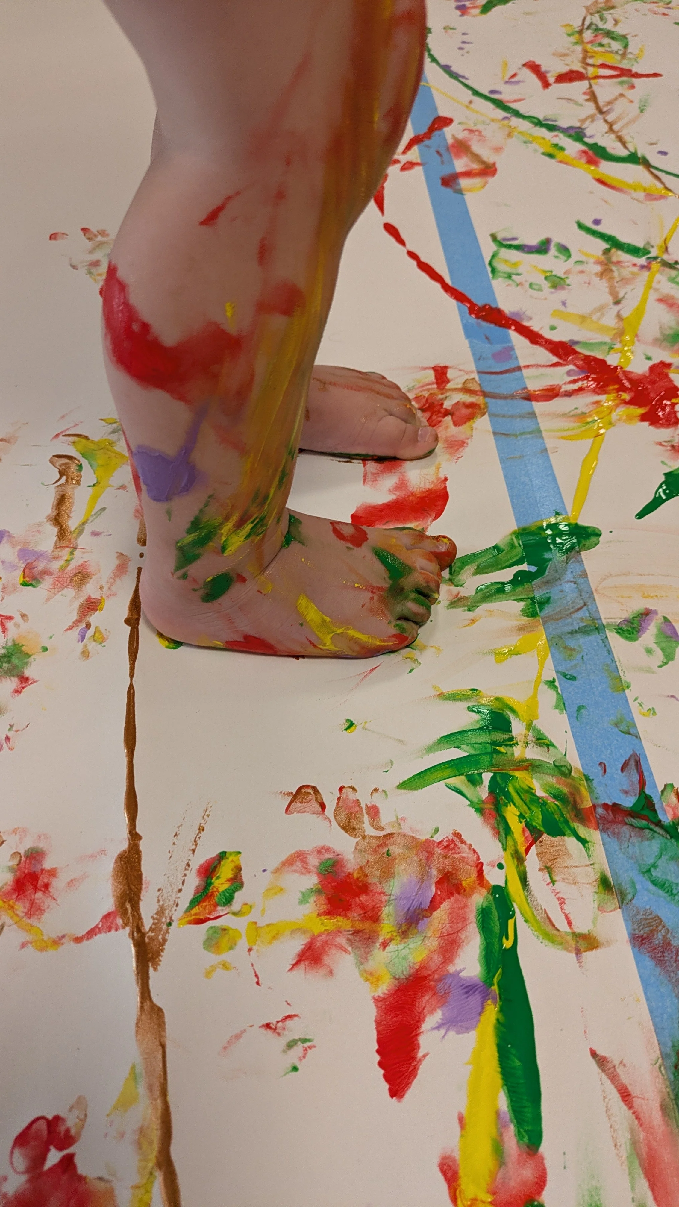 Et barn står barfotet på et stort papir eller hvit duk, med maling både på papiret og oppover beina