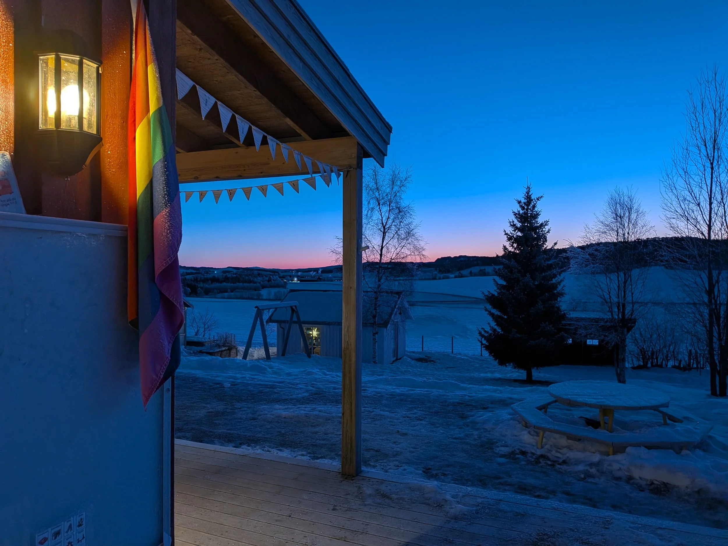 Utendørsbilde av barnehagen vinterstid, med en rosa og blå himmel i bakgrunnen. Pride-flagget henger på veggen av bygningen
