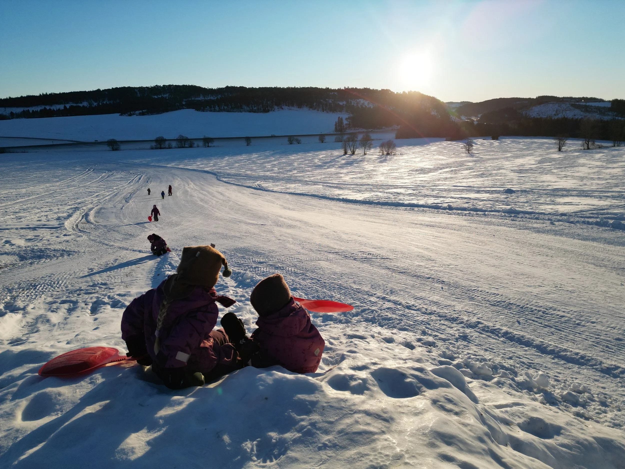 Barn som sitter i snøen på toppen av en akebakke. I bakgrunnen snødekt landskap, åser og blå himmel med sol