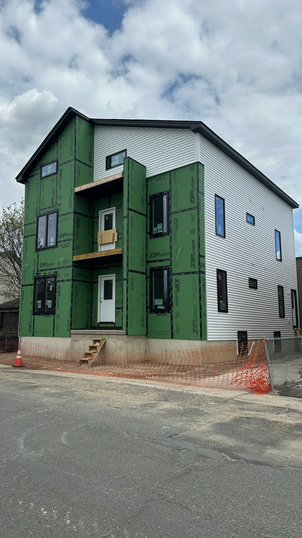 Under construction multi-story house with green insulation, white siding, and black window frames, surrounded by an orange safety fence.