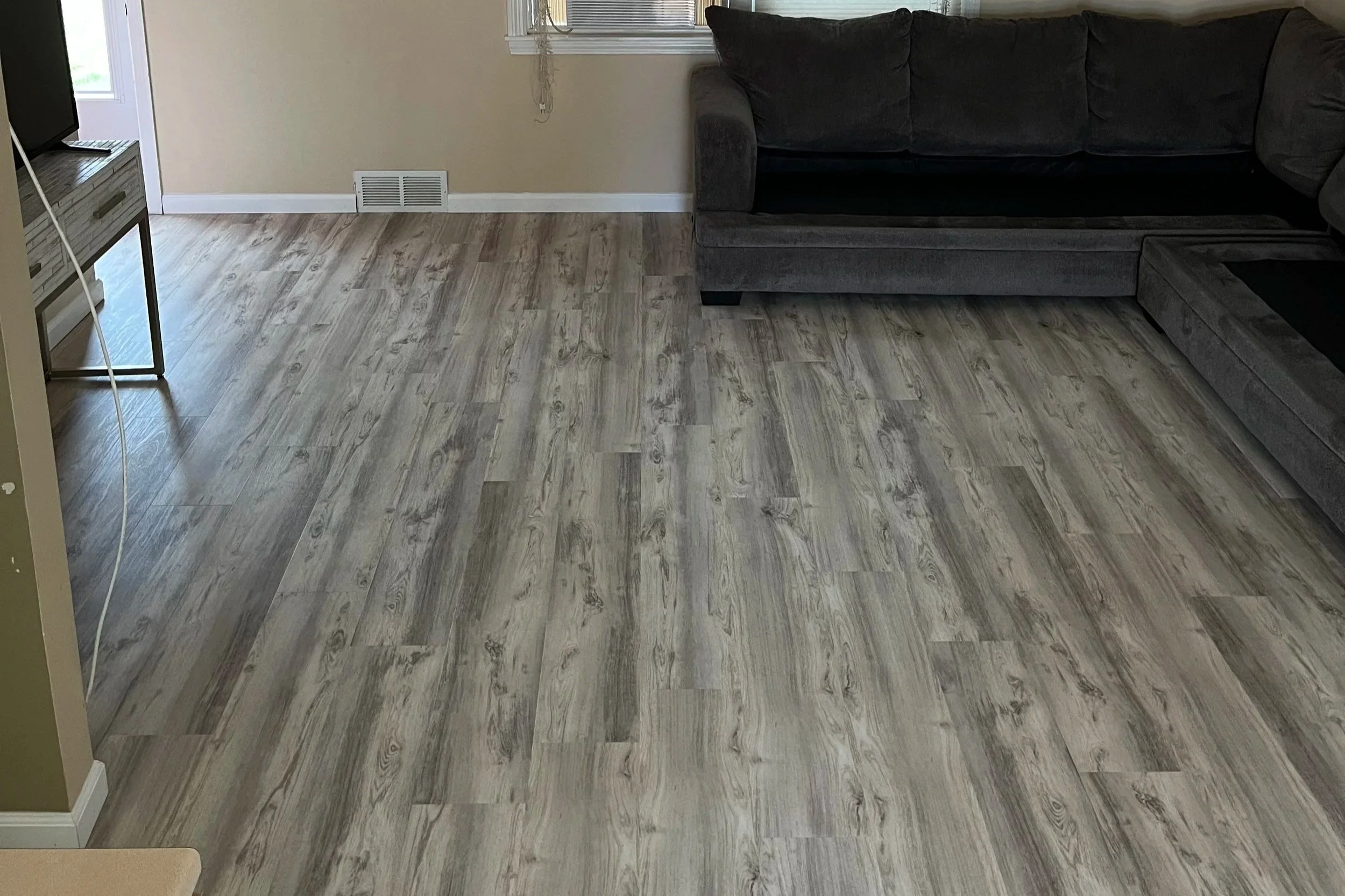 Living room with wood-look flooring, a black sectional sofa, beige walls, and a window with blinds.
