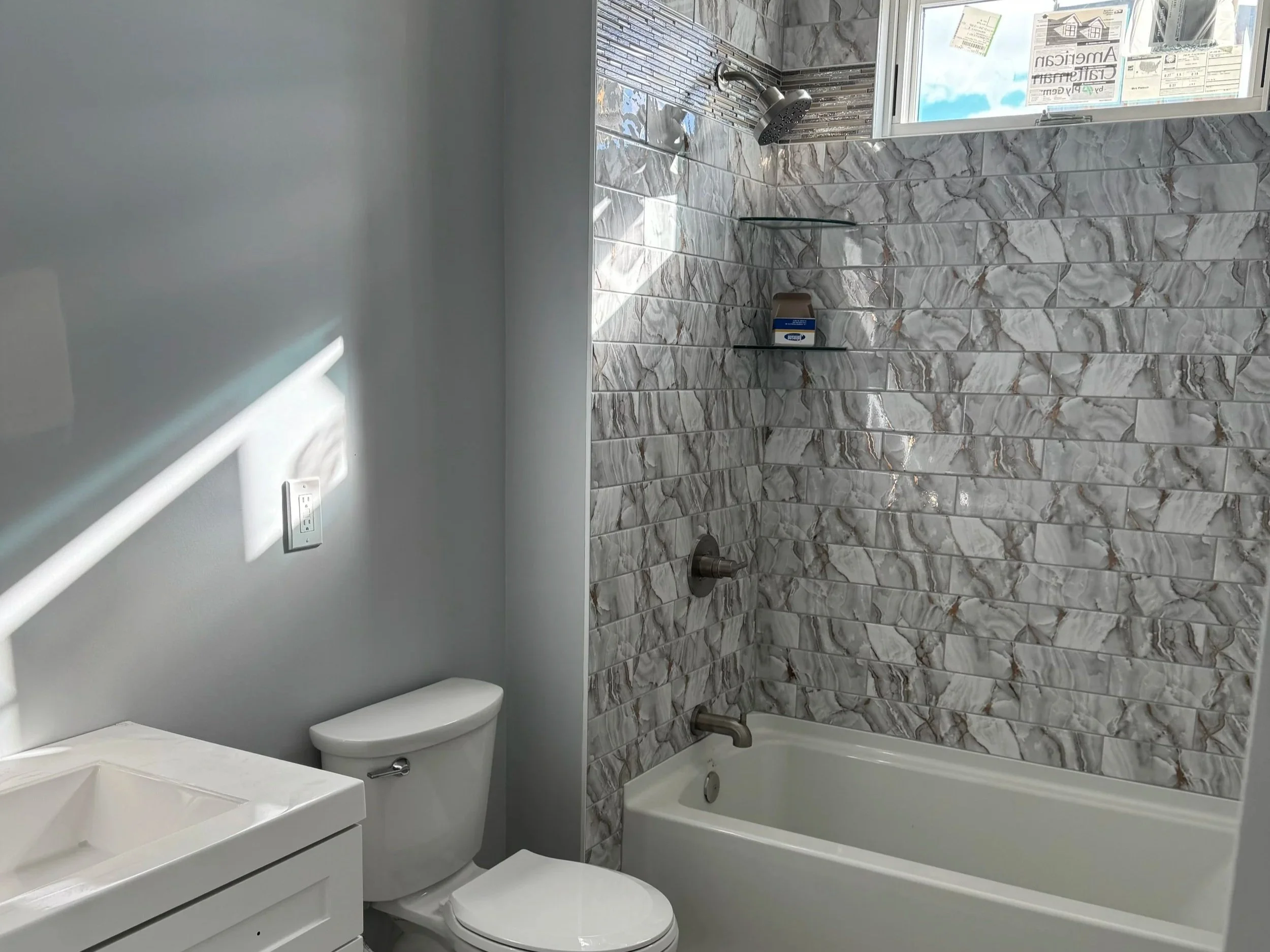 Bathroom with a bathtub and shower combination with gray and white marble tile walls, a small window, a showerhead, and two glass shelves. There is also a white toilet and a sink vanity, with a wall outlet and a light-colored wall.