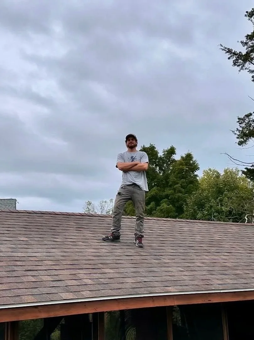 A man standing on a roof with crossed arms, wearing a gray t-shirt, gray pants, and a cap, with trees and cloudy sky in the background.