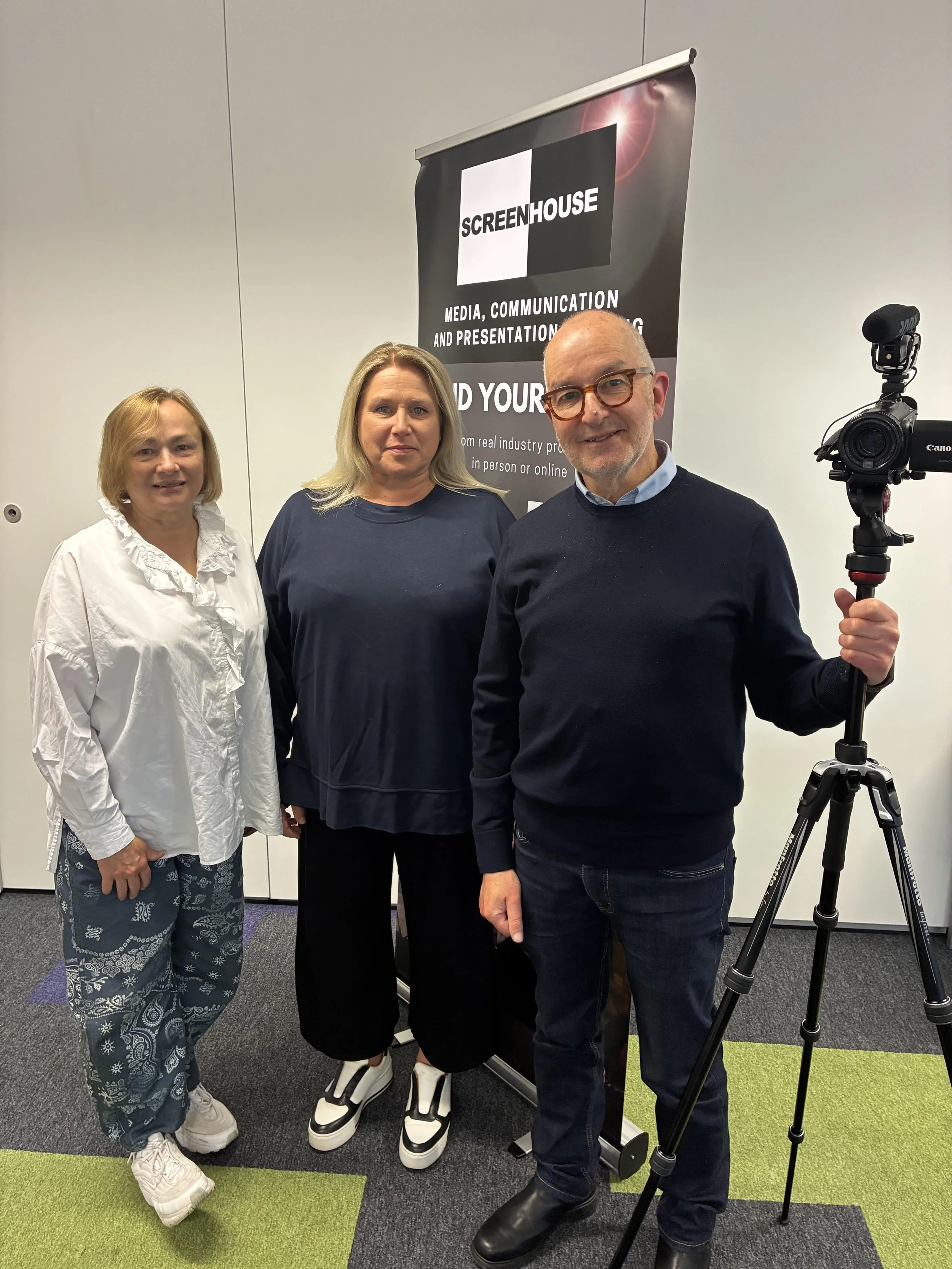 Three people standing indoors, with a banner in the background that says 'SCREENHOUSE MEDIA, COMMUNICATION AND PRESENTATION TRAINING'. The person on the right is holding a professional video camera on a tripod.