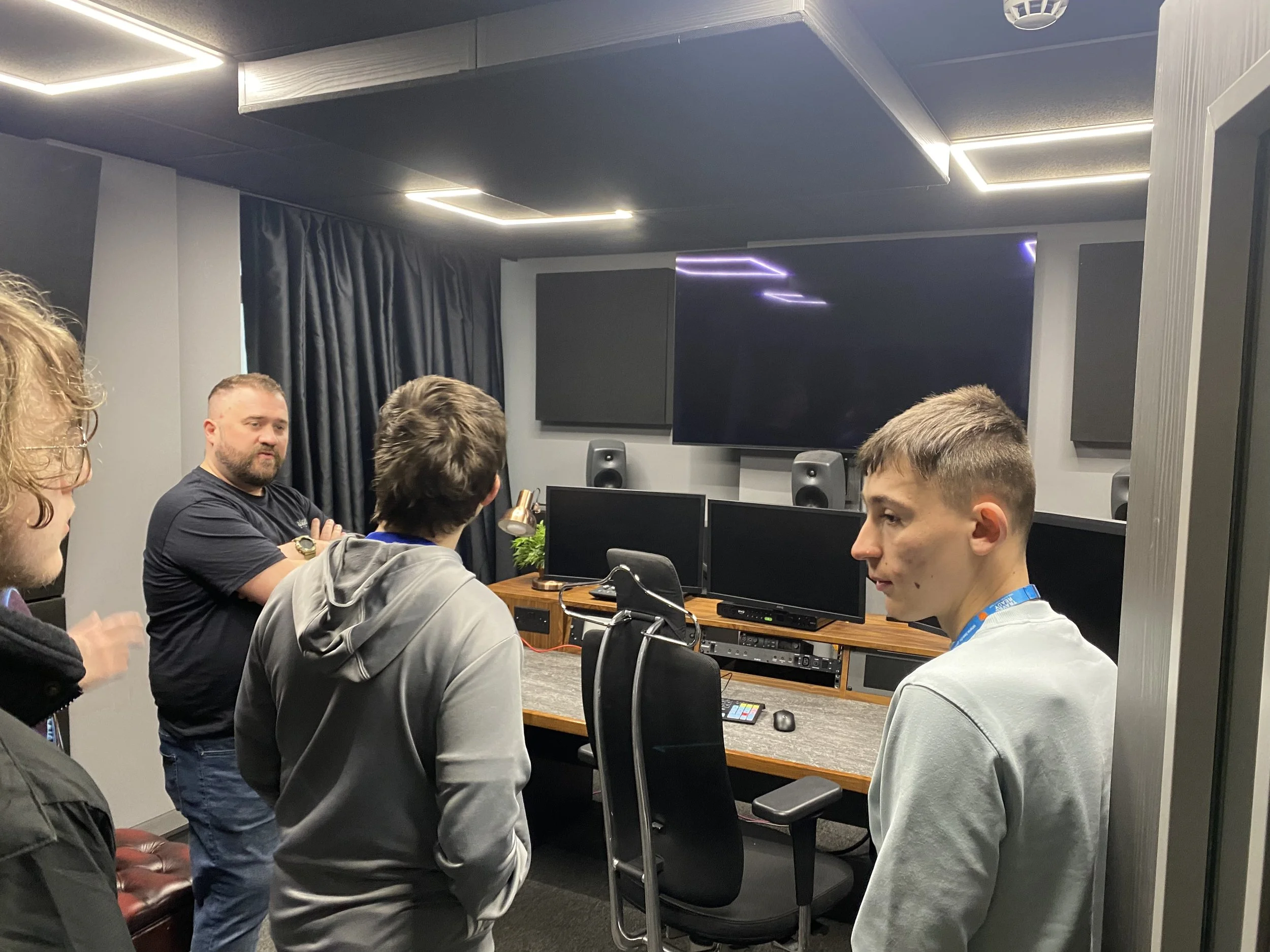 A group of young men having a discussion in a modern recording studio, with digital equipment, monitors, and a large screen in the background.