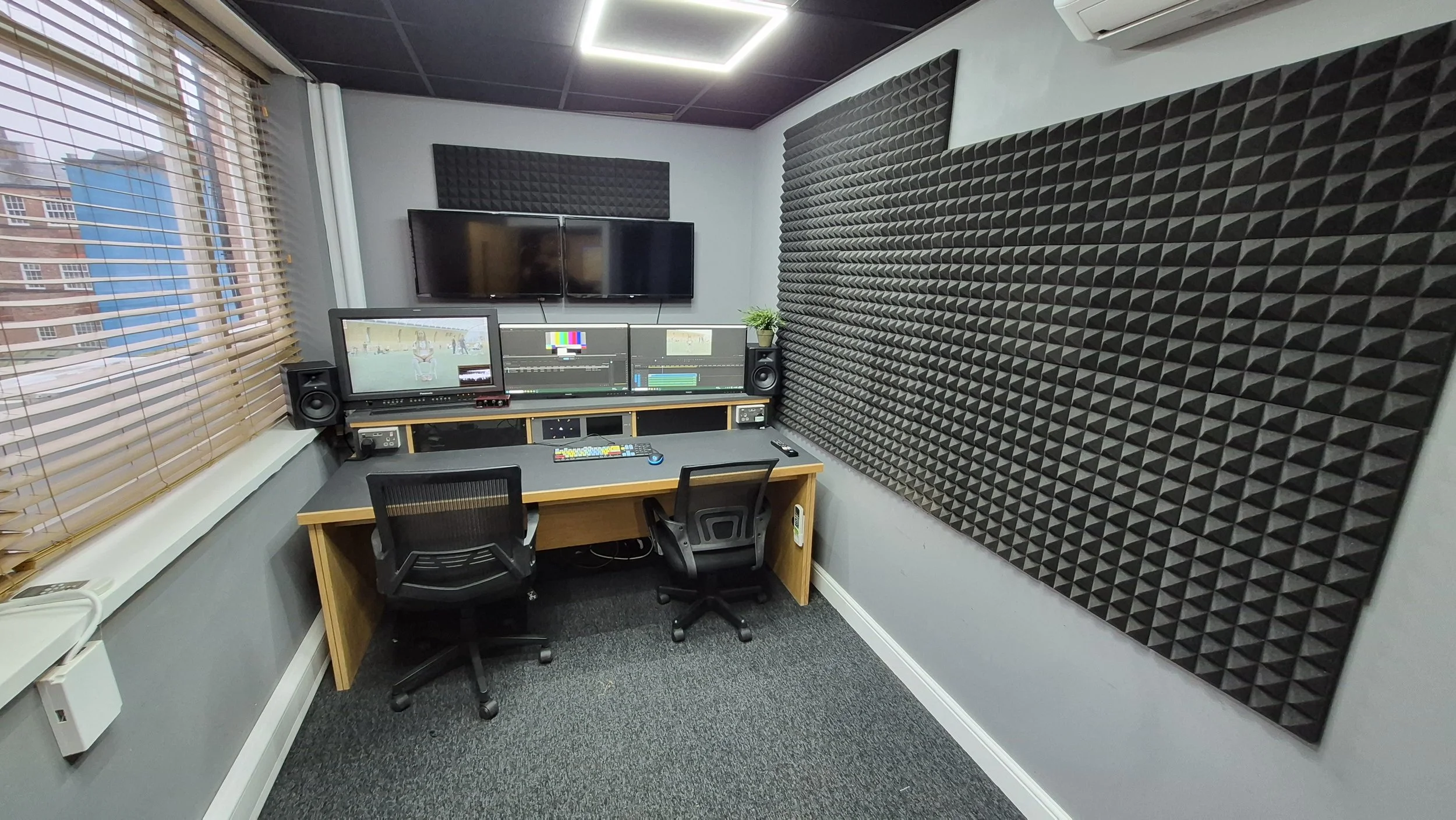 A control room with two computer monitors, video editing equipment, studio speakers, and acoustic foam on the walls, with large windows on the left side showing city buildings outside.