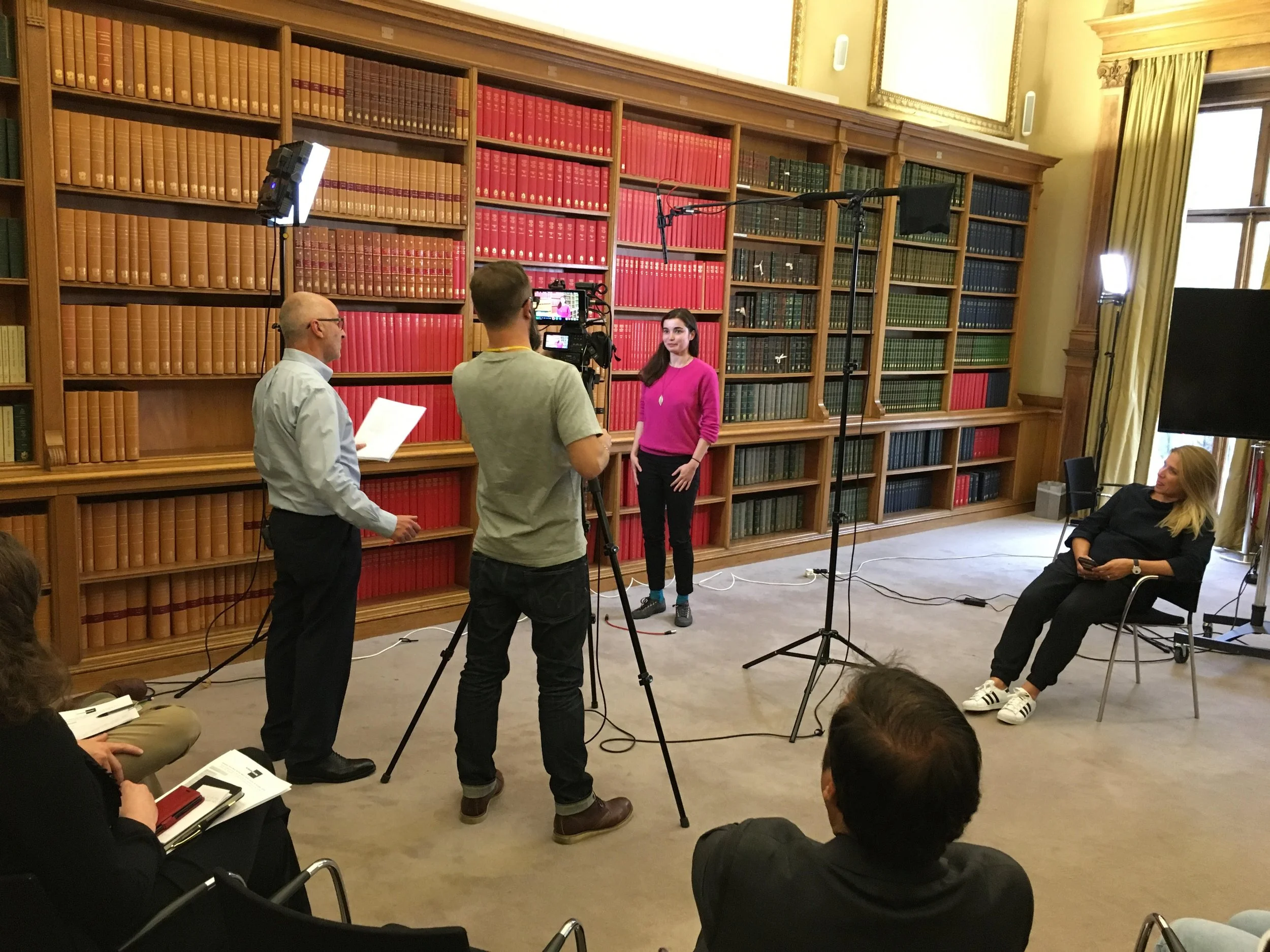 A video interview or recording session in a library with bookshelf background, featuring a woman in a pink sweater being filmed by a crew. Crew members are handling camera and lighting equipment, and others are seated, listening, with notebooks.