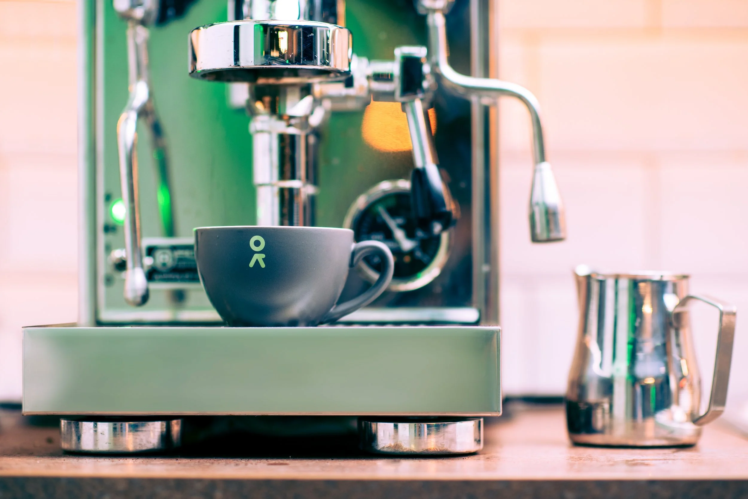 A modern stainless steel espresso machine brewing coffee into a black cup on a wooden countertop, with a shiny milk frothing pitcher to the right.