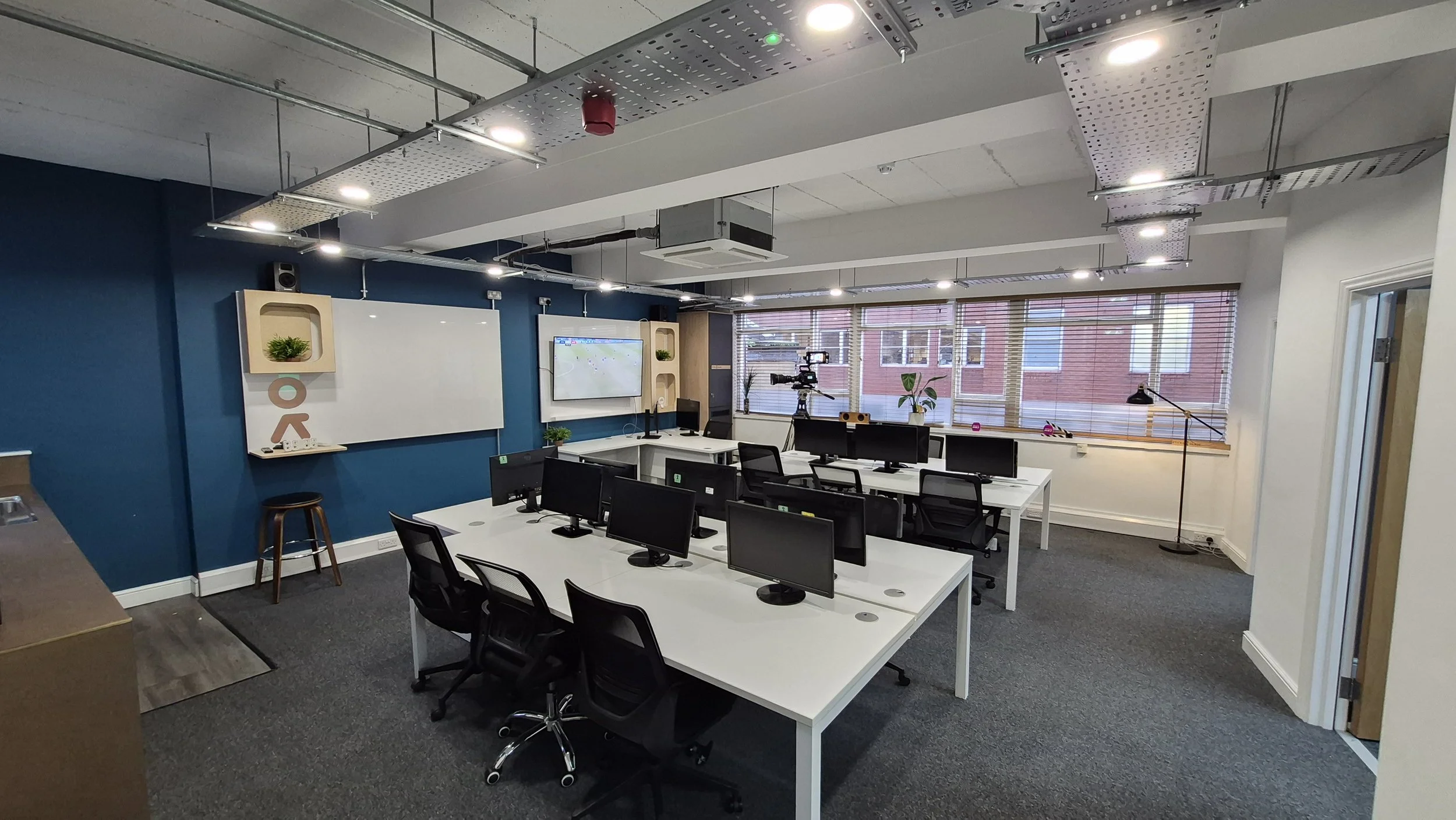 Modern office classroom with white desks, multiple computer monitors, black chairs, a whiteboard, a large window with blinds, some plants, and a television screen on a blue accent wall.