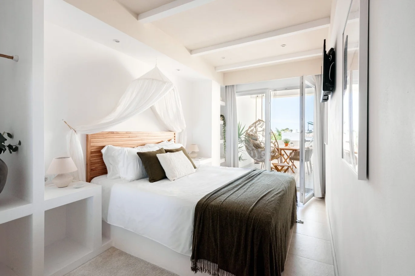 Bright bedroom with a wooden bed frame, white bedding, and multiple pillows, with a balcony view of outdoor furniture and plants. Holiday home in Lagos Algarve
