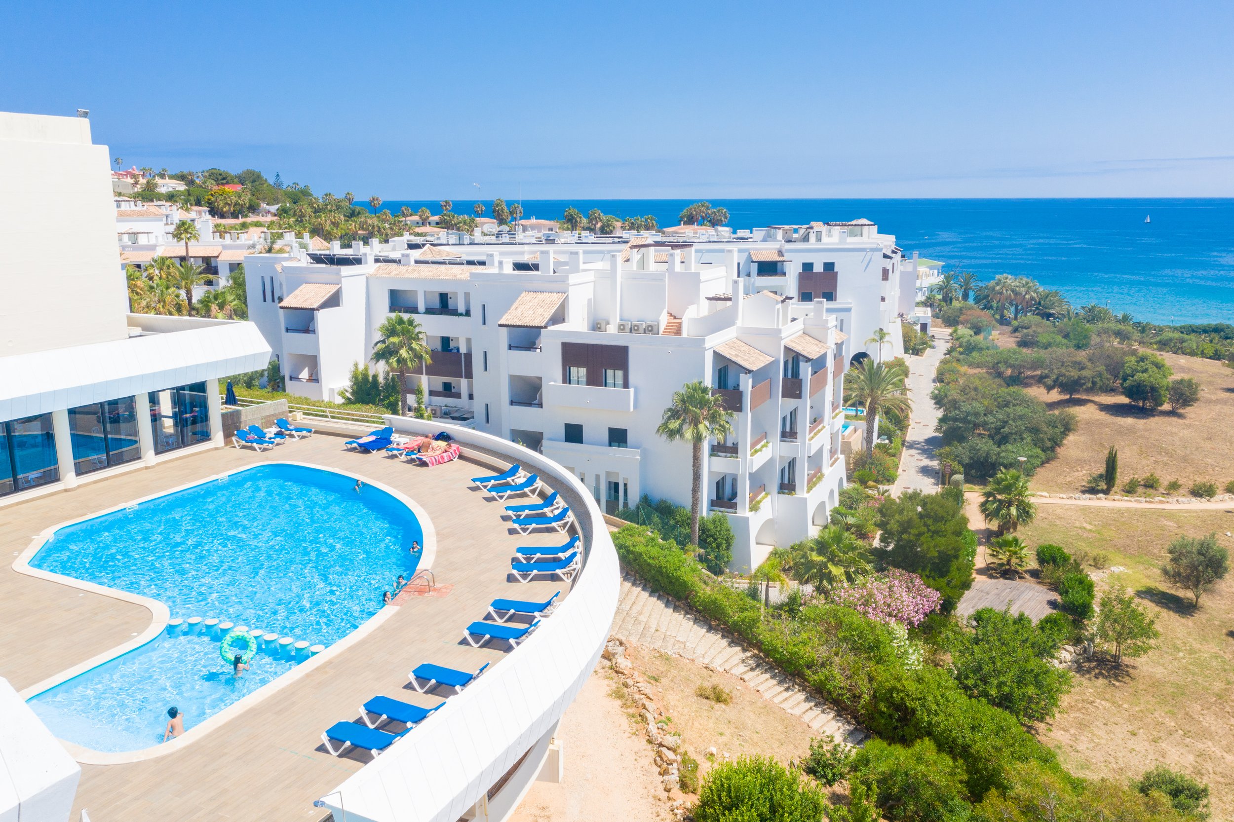 Aerial view of a modern white apartment complex with a curved swimming pool surrounded by blue lounge chairs and palm trees, overlooking the ocean. Holiday home in Lagos Algarve