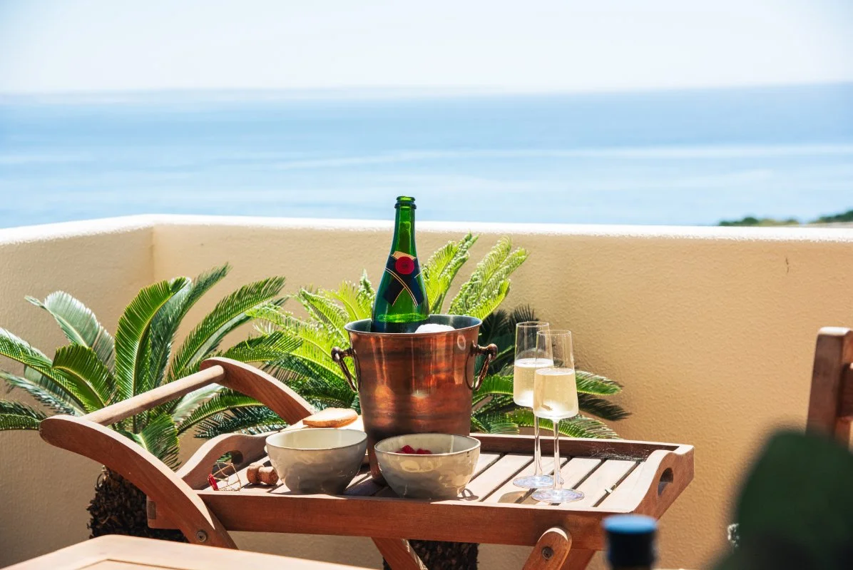 A table with two glasses of champagne, bowls, and a bottle of sparkling wine or champagne in an ice bucket, set on a balcony with green plants and a view of the ocean in the background.