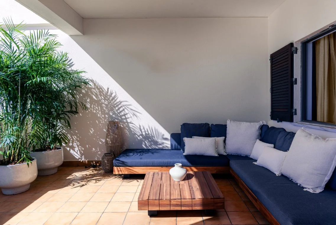 Outdoor patio area with a built-in L-shaped blue cushioned sofa, white pillows, a wooden coffee table with a white vase, large potted green plants, and a wall with a window and black shutters, illuminated by sunlight.