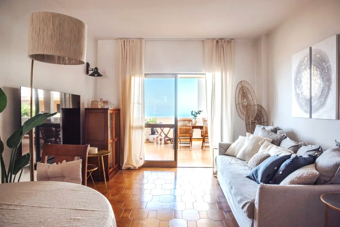 Living room with a sofa, dining area on balcony, ocean view through glass sliding door, wooden furniture, decorative pillows, artwork on wall, natural light.