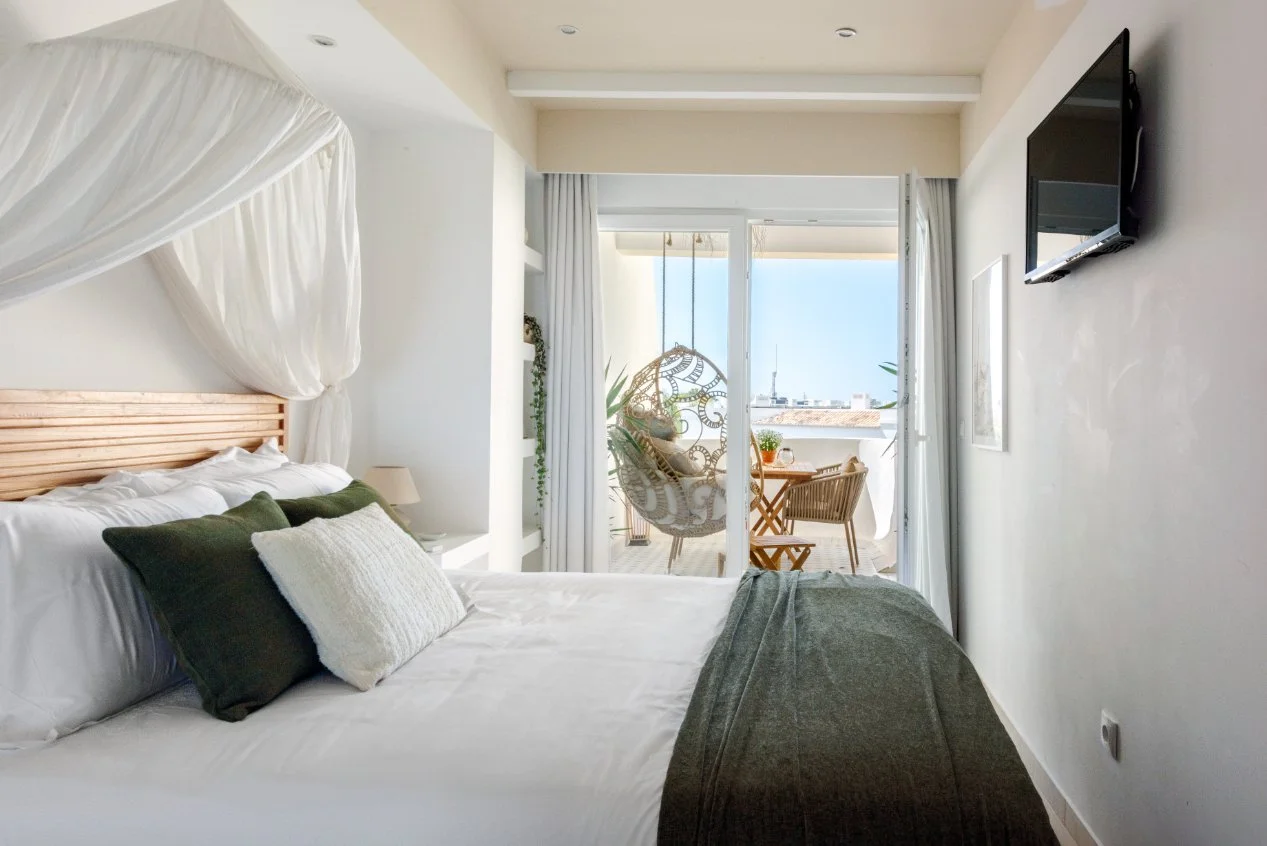 A cozy bedroom with a large bed featuring white linens, dark green and cream pillows, and a wooden headboard. A canopy or curtain drapes over part of the bed. The room opens to a balcony with a hanging chair, small table, and two chairs, overlooking 