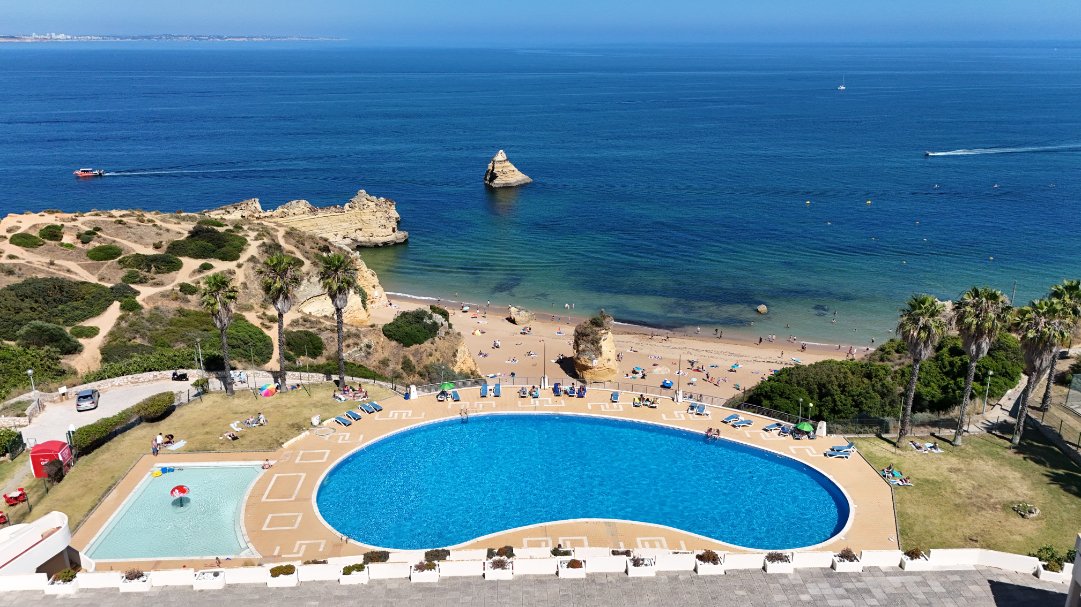 A scenic view of Lagos beach with sunbathers, turquoise water, and a large rock formation in the ocean. In the foreground, a swimming pool and smaller pool are visible, surrounded by lounge chairs, palm trees, and grassy areas.