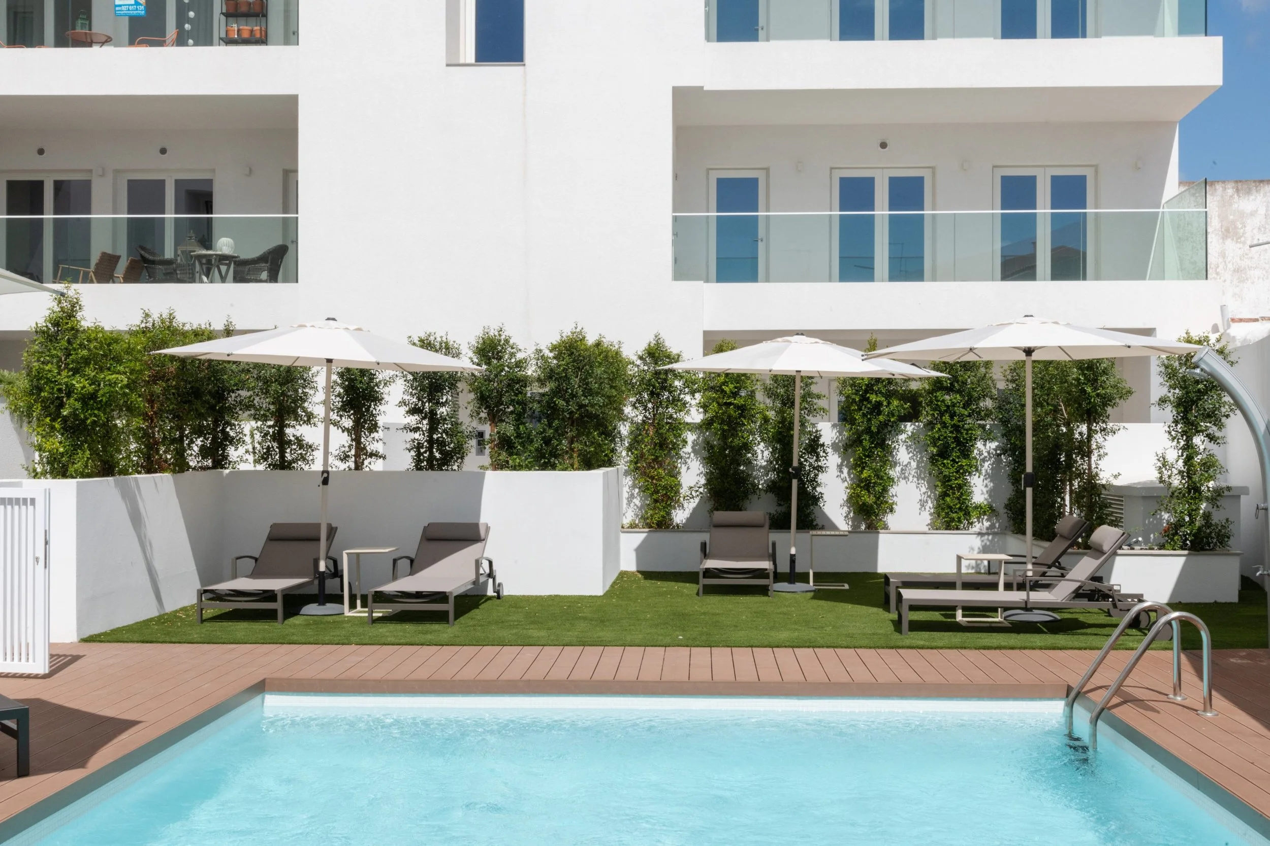 A swimming pool in the foreground with a blue water surface, surrounded by a wooden deck. In the background, there are lounge chairs with umbrellas, a grassy area. Newly built rental apartment in Lagos near the beach.