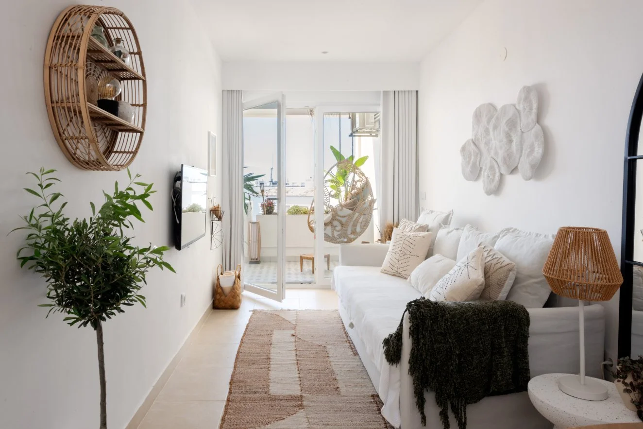 A living room with white walls and a white couch decorated with beige pillows, a green throw blanket, and a wall-mounted white art piece made of fabric. There is a wicker side table with a wicker lamp next to the couch. A glass door leads to a balcony with potted plants, a hanging bamboo chair, and a view of a harbor or waterfront outside.