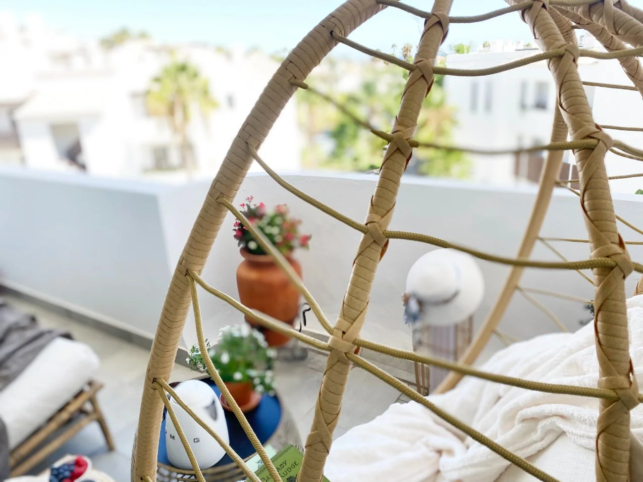 Balcony with wicker hanging chair, potted flowers, and a white blanket, overlooking trees and apartment buildings. Holiday home in Lagos Algarve
