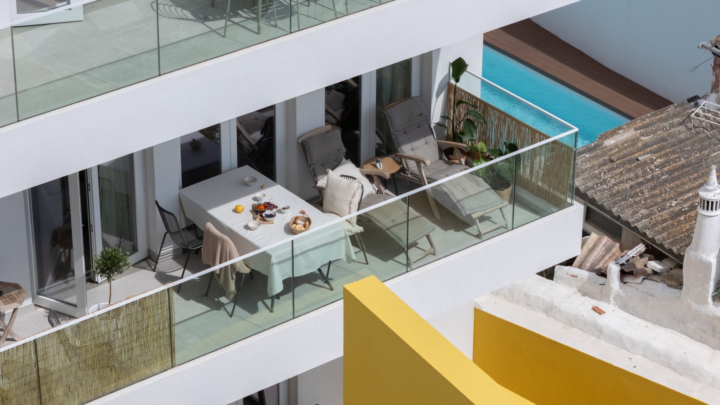 Balcony with outdoor dining table, chairs, lounge chairs, potted plants, and swimming pool on a modern building. Terraced rental apartment in Tavira near the beach.