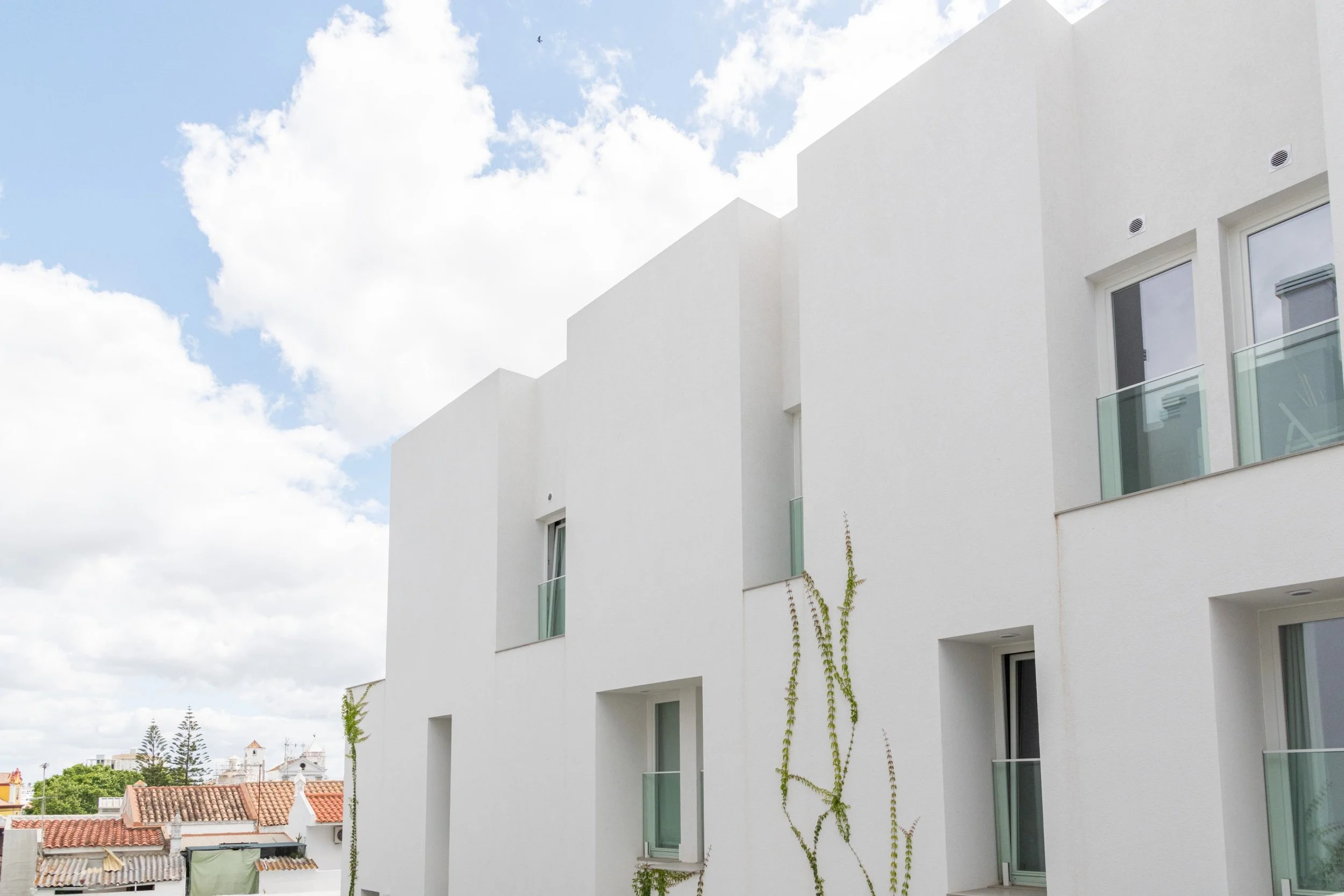 White modern apartment building with glass balconies and sliding doors, partly visible sky with clouds in the background.