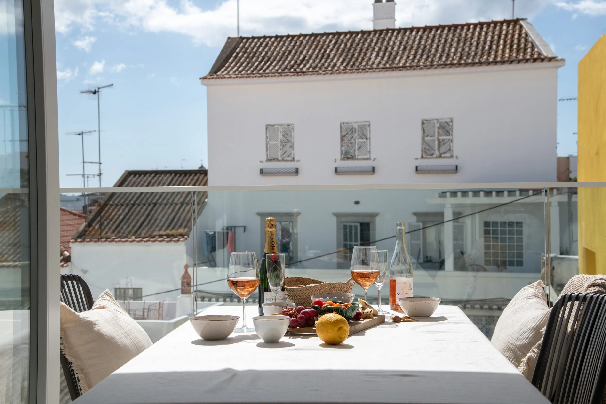 Outdoor dining table with wine glasses, a bottle of rosé, a bowl with grapes, apple, lemon, and bread on a balcony with a city view. Terraced rental apartment in Tavira near the beach.