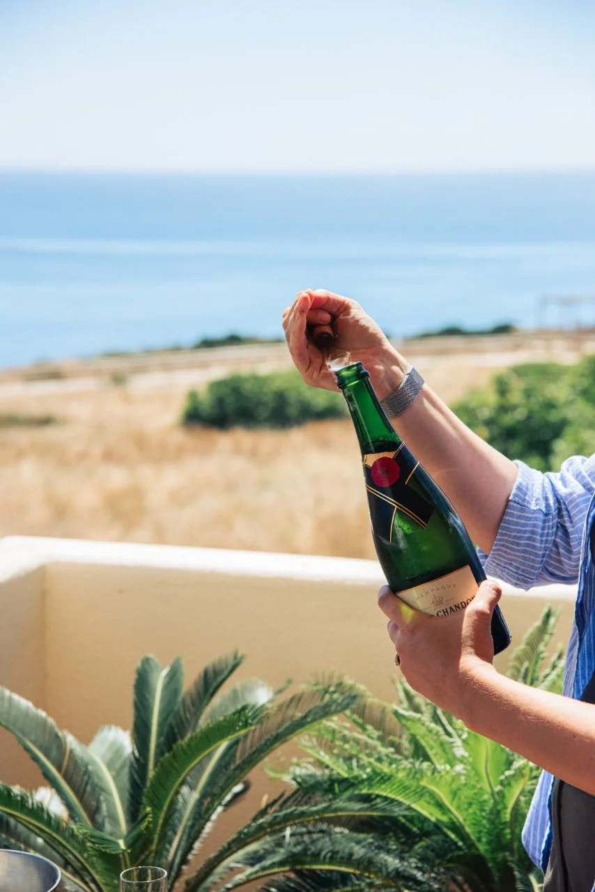 Person opening a bottle of champagne on a balcony overlooking the ocean with greenery in the foreground.
