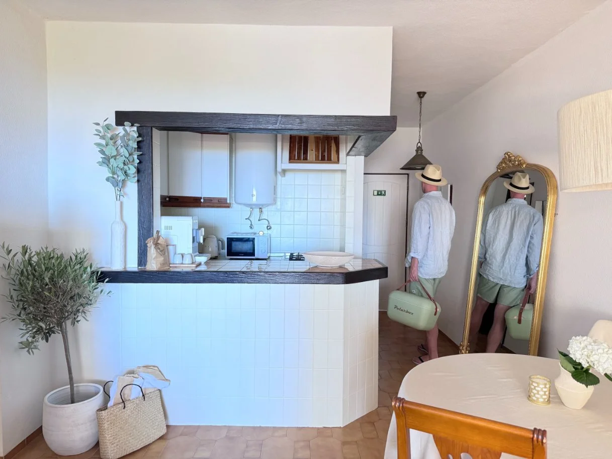 A man in a white shirt, shorts, and a straw hat standing in a kitchen, holding a pet carrier, is reflected in a full-length mirror. The kitchen has white walls, a black wood trim, and various appliances on the counter.  Holiday home in Lagos Algarve