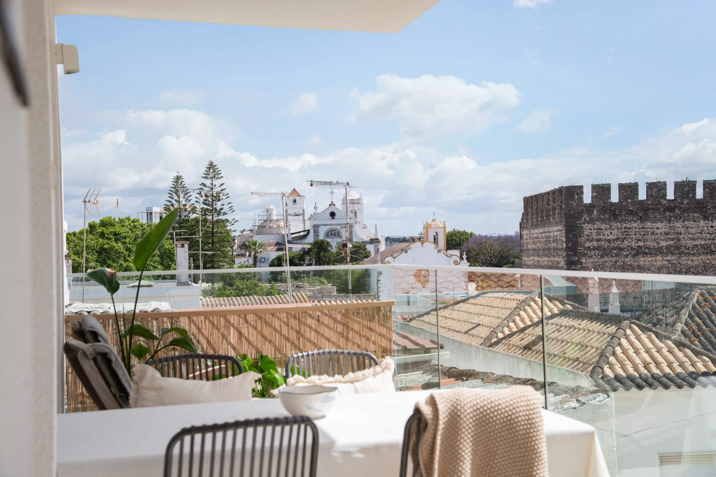 Balcony with a table, chairs, and cushions overlooking rooftops and a historic church with bell towers, trees, and a large stone wall in the background on a sunny day.