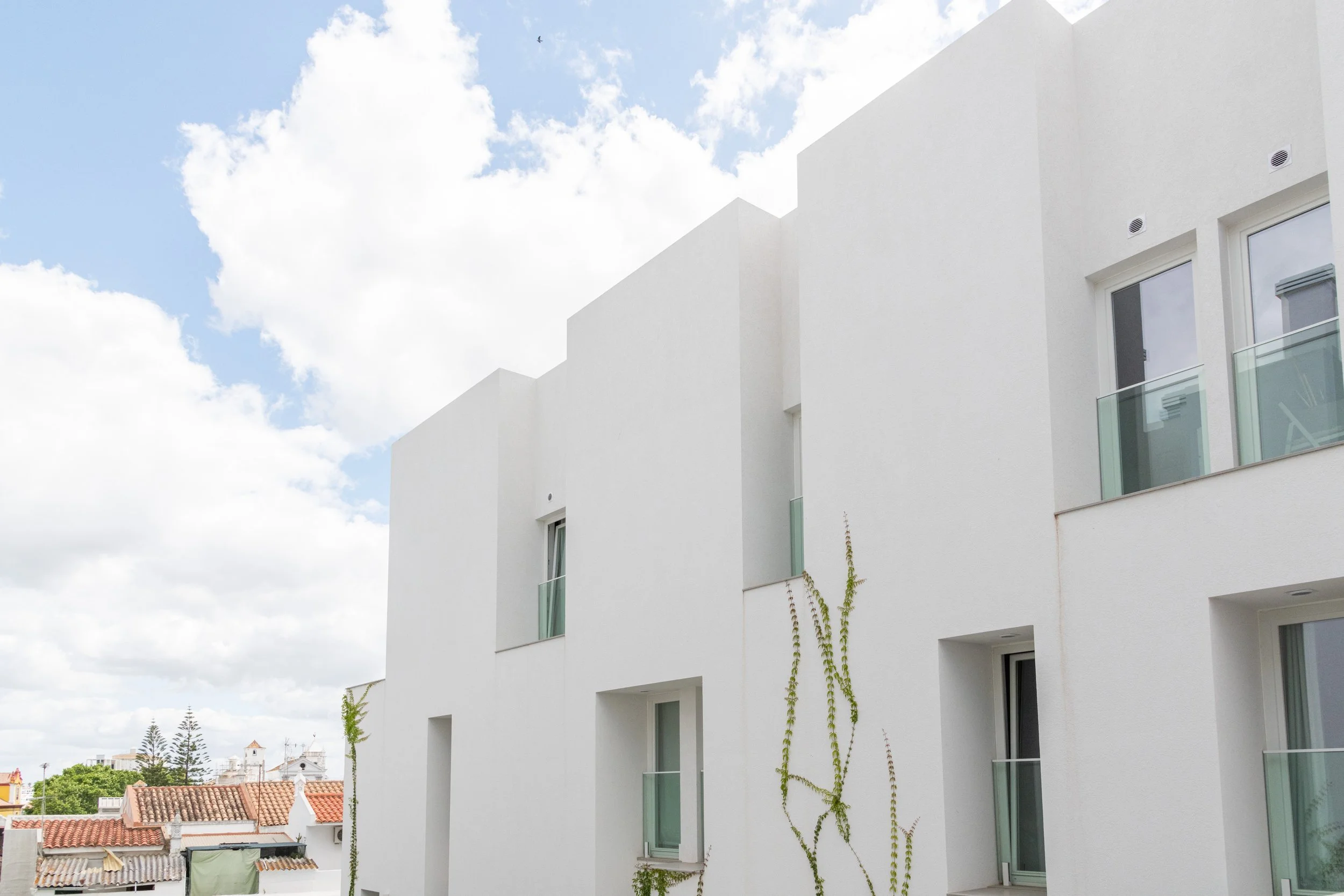 White modern apartment building with glass balconies and a few climbing vines, under a partly cloudy sky.