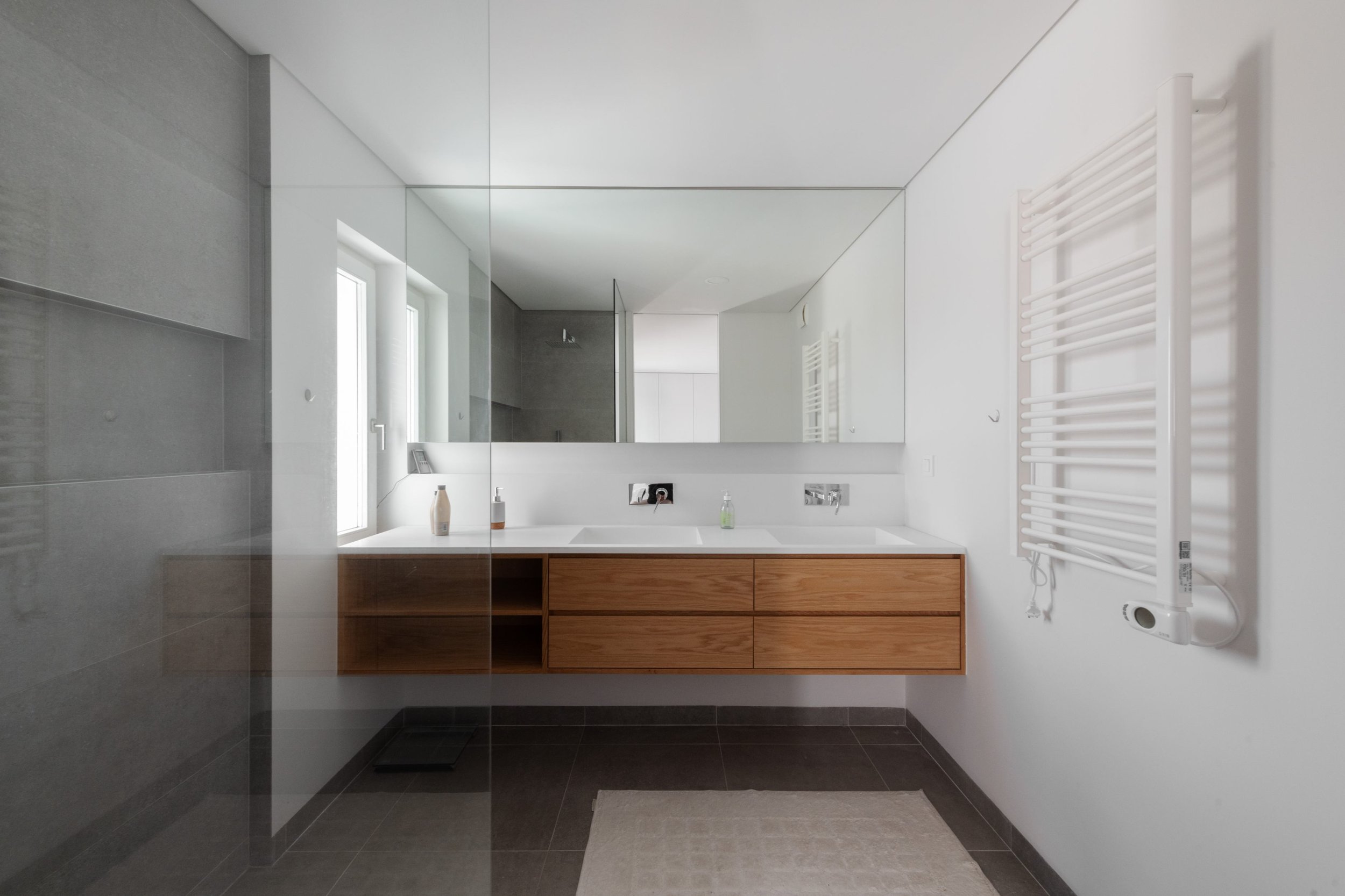 Modern bathroom with a large mirror, white walls, wooden vanity, black floor tiles, and a white towel radiator on the right.
