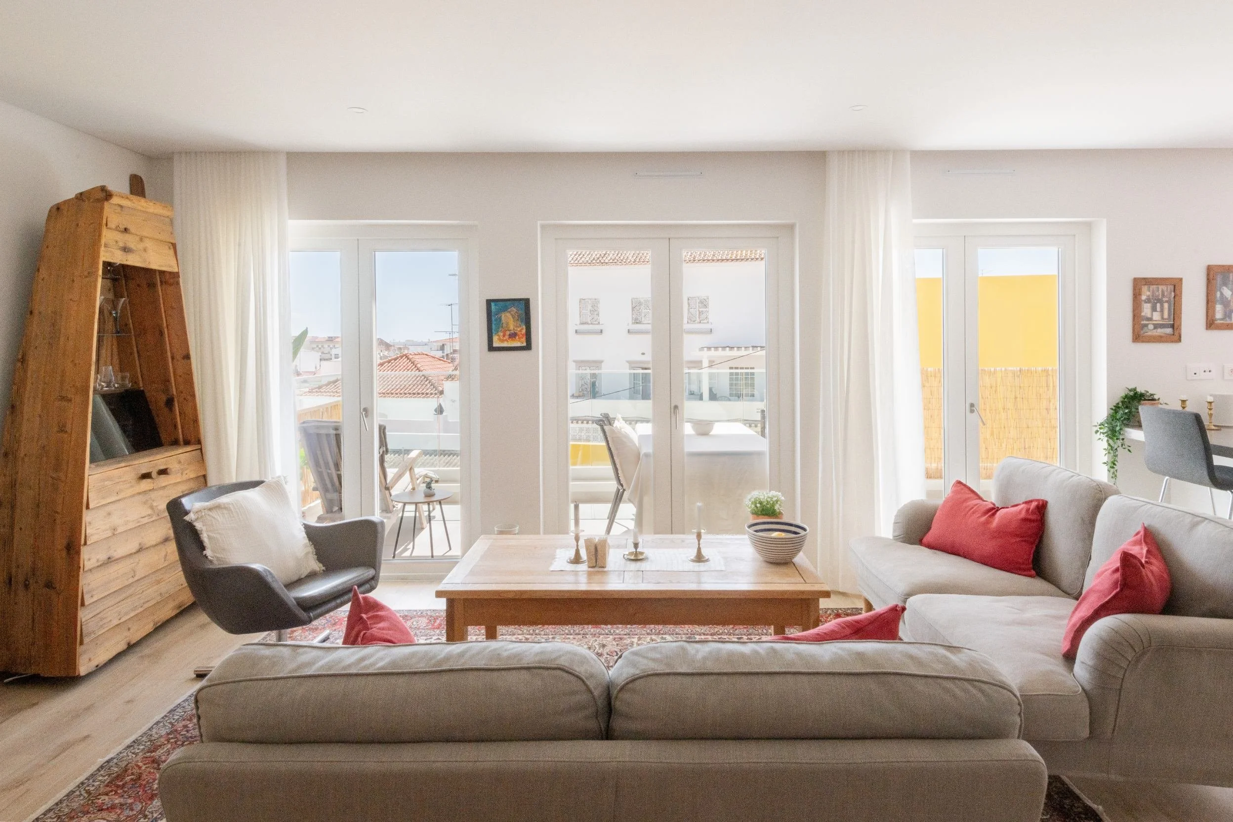 Bright living room with large glass doors leading to a balcony, featuring beige sofas with red pillows, a black armchair, a wooden coffee table, and a wooden bookshelf on the left, with white curtains and outdoor furniture visible through the doors.