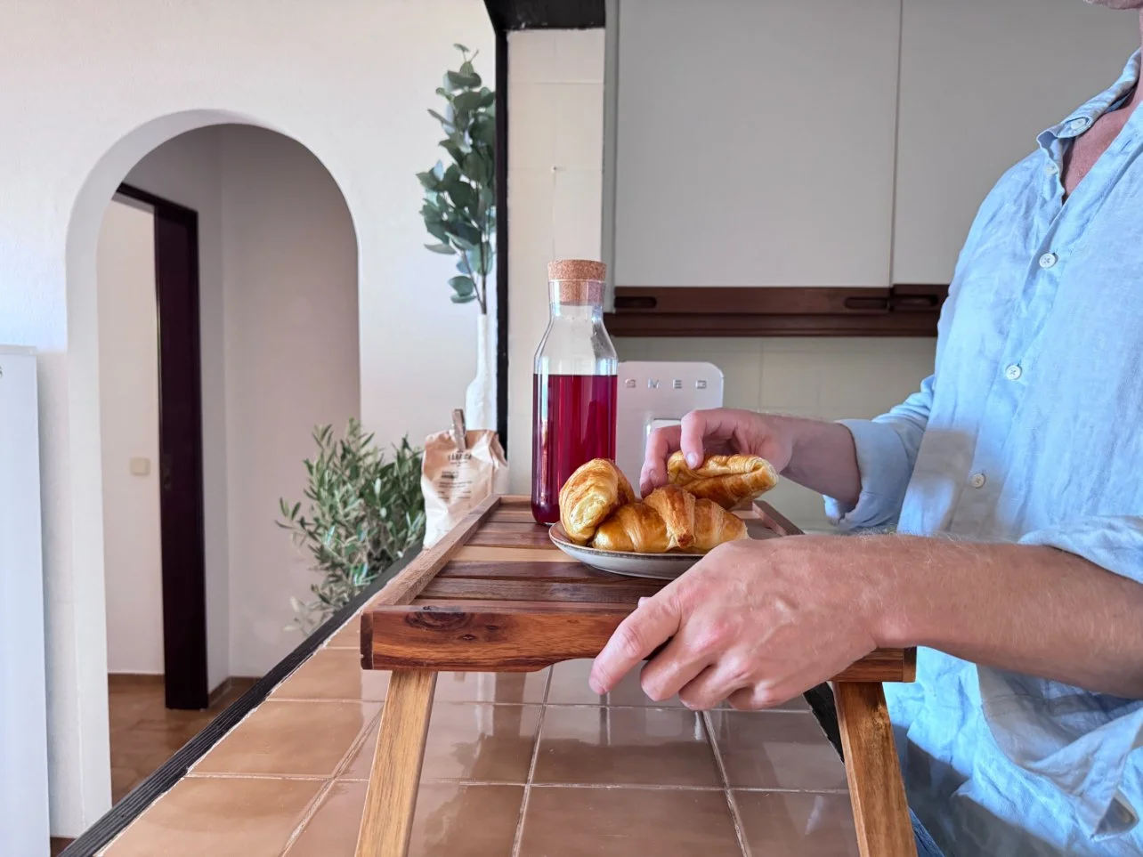Person reaching for croissants and a glass of red juice on a wooden tray, with a kitchen background. Holiday home in Lagos Algarve