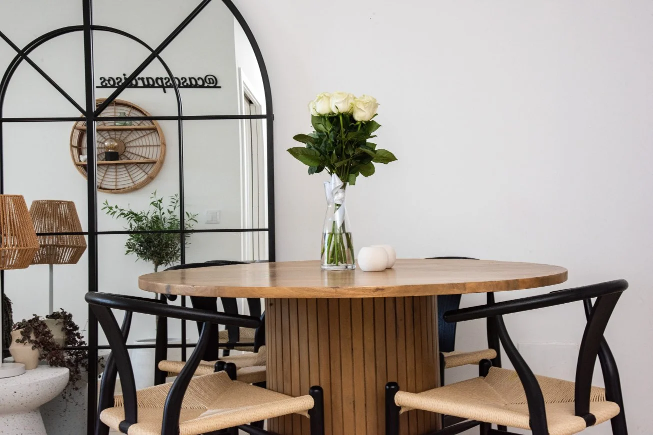 A round wooden table with a glass vase of white roses and two small white candles, surrounded by black chairs with woven seats, in a modern dining room.