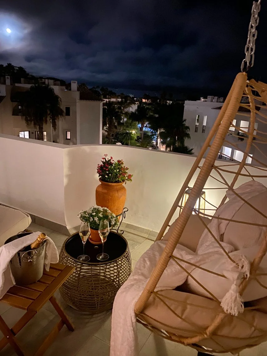 A cozy balcony at night with two potted plants, a small table with two glasses of champagne, a lounge chair, and a decorative hanging chair. The background shows a dark, cloudy sky. Holiday home in Lagos Algarve.