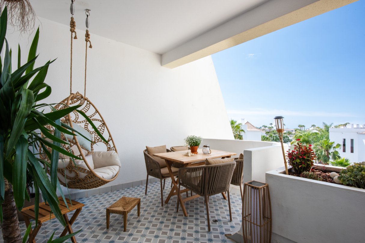 Outdoor patio with a hanging wicker egg chair, a wooden dining table with six chairs, potted plants, and a view of a coastal neighborhood with palm trees under a clear blue sky.