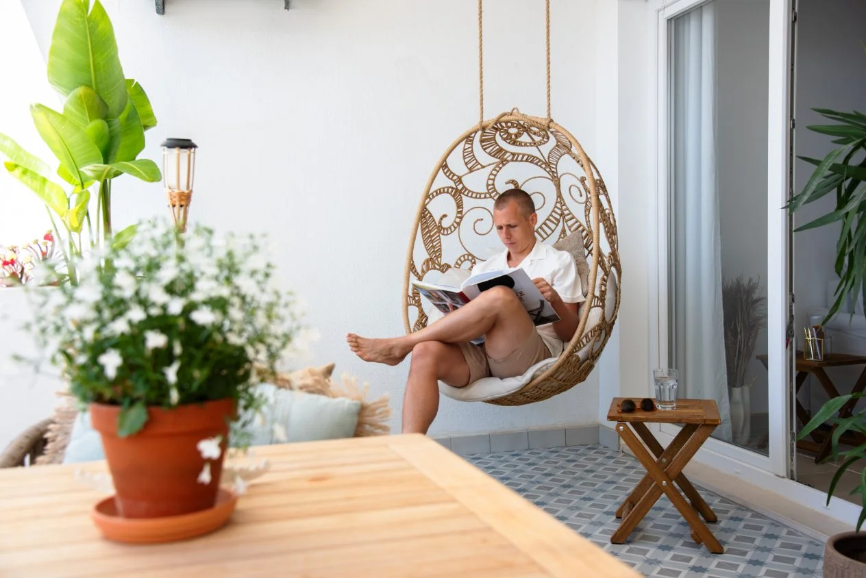 A man sitting on a hanging wicker chair on a balcony, reading a magazine. There are plants, a wooden table, a glass of water, and a cat in the background.