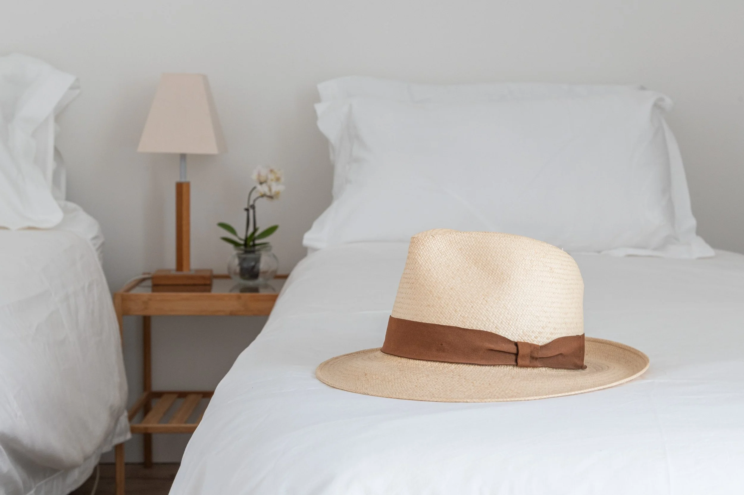 A straw sun hat with a brown ribbon resting on a white bed in a minimalist bedroom with a wooden nightstand, lamp, and orchid plant.