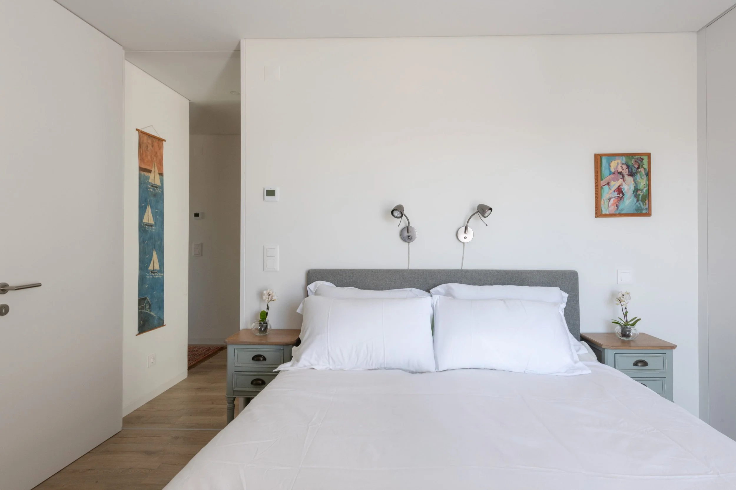 A bedroom with a bed, gray headboard, white bedding, and two pillows. Two nightstands each with a small glass vase with a flower. Two wall-mounted reading lamps above the bed. Two paintings on the white walls, one with sailboats and the other with a 