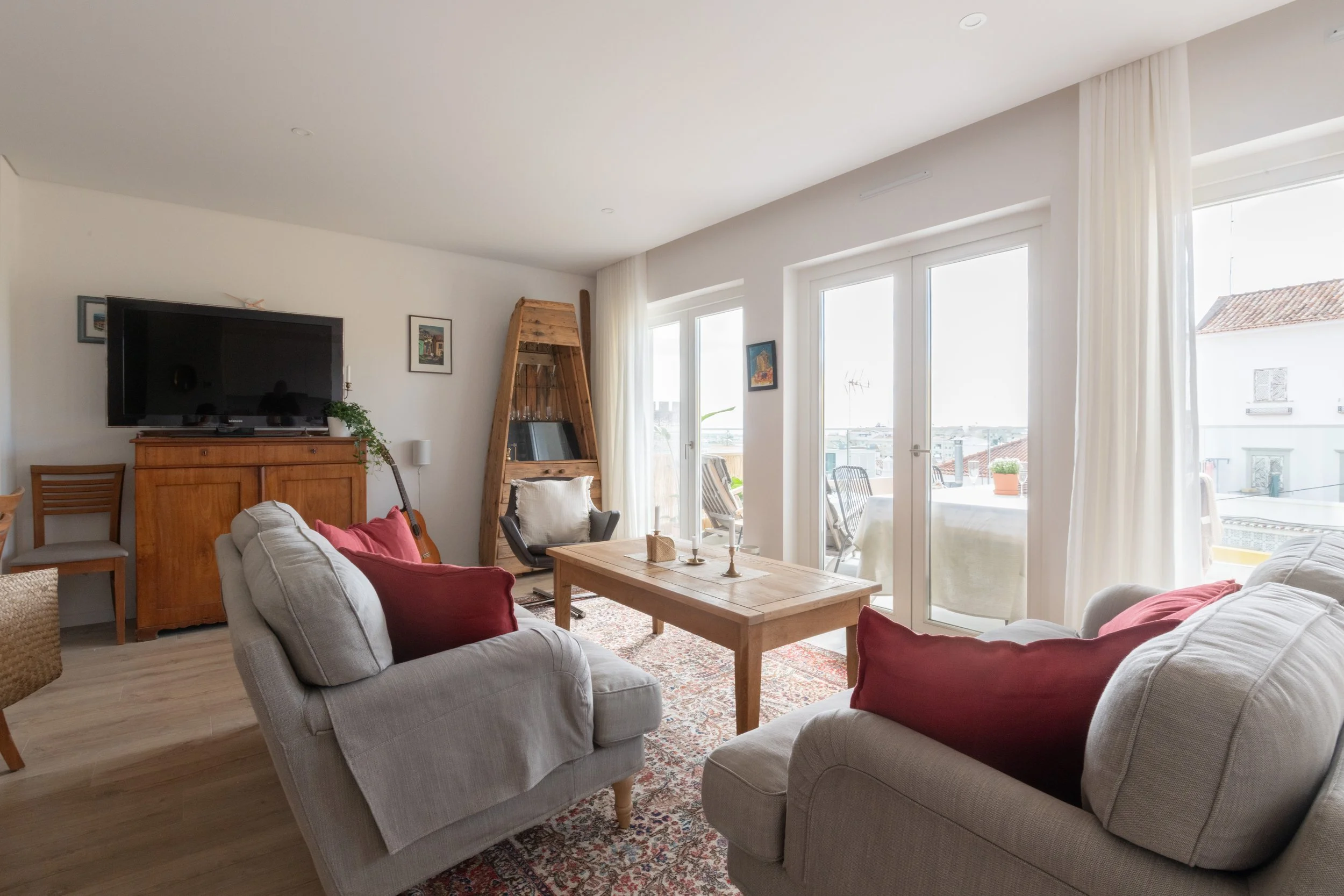 Living room with two beige sofas, red pillows, a wooden coffee table, a large flat-screen TV on a wooden cabinet, a wooden bookcase, and glass patio doors leading to a balcony with outdoor chairs and potted plants, bright natural light.