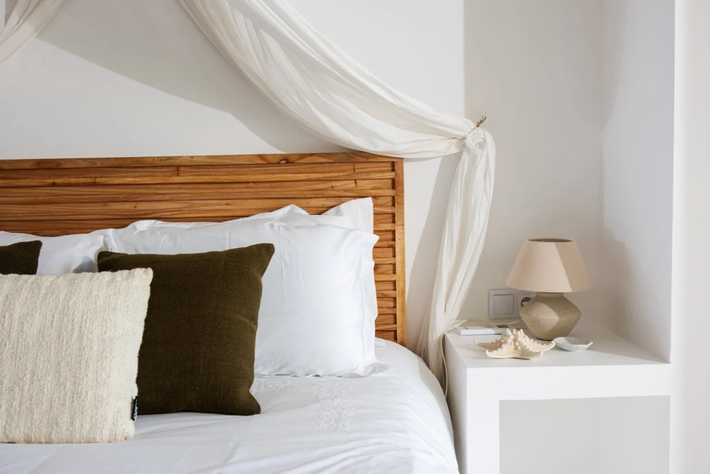 A neatly made bed with white sheets, three pillows in dark and light colors, a wooden headboard, a small white bedside table with a beige lamp, a decorative shell, and a surfing board. A white curtain is draped over the headboard.
