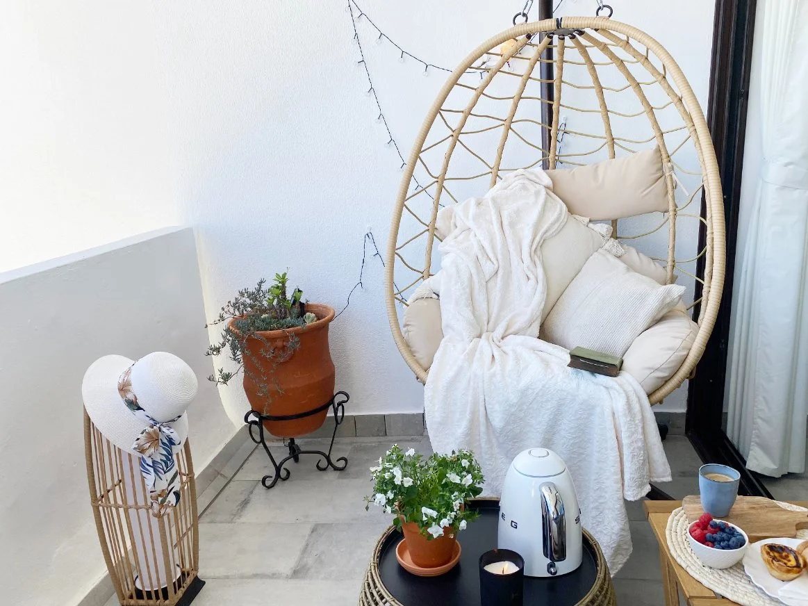 Cozy balcony corner with a hanging wicker egg chair draped in a blanket and pillows, a small table with a potted plant, a kettle, a candle, a cup of coffee. Holiday home in Lagos Algarve.
