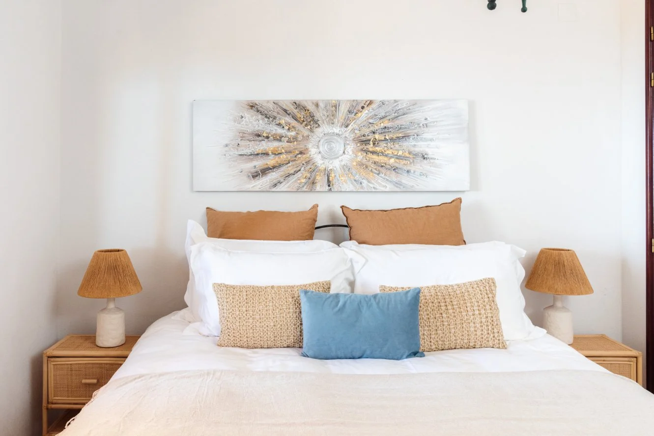 A neatly made bed with four pillows and a white quilt, flanked by two wooden nightstands with matching lamps; a white wall with an abstract painting hangs above the bed. Holiday home in Lagos Algarve