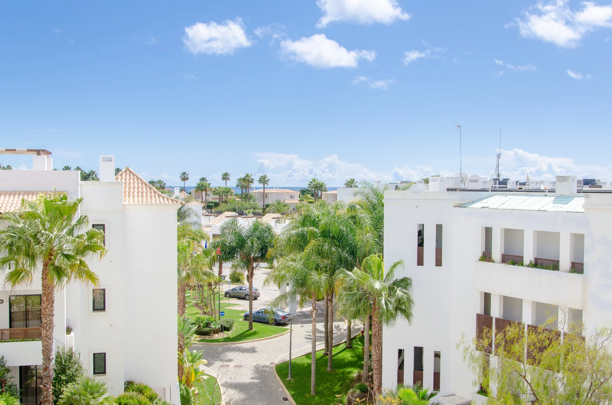 A bright, sunny day in a modern city neighborhood with white residential buildings, palm trees, a parking area, and a blue sky with some clouds. Holiday home in Lagos Algarve