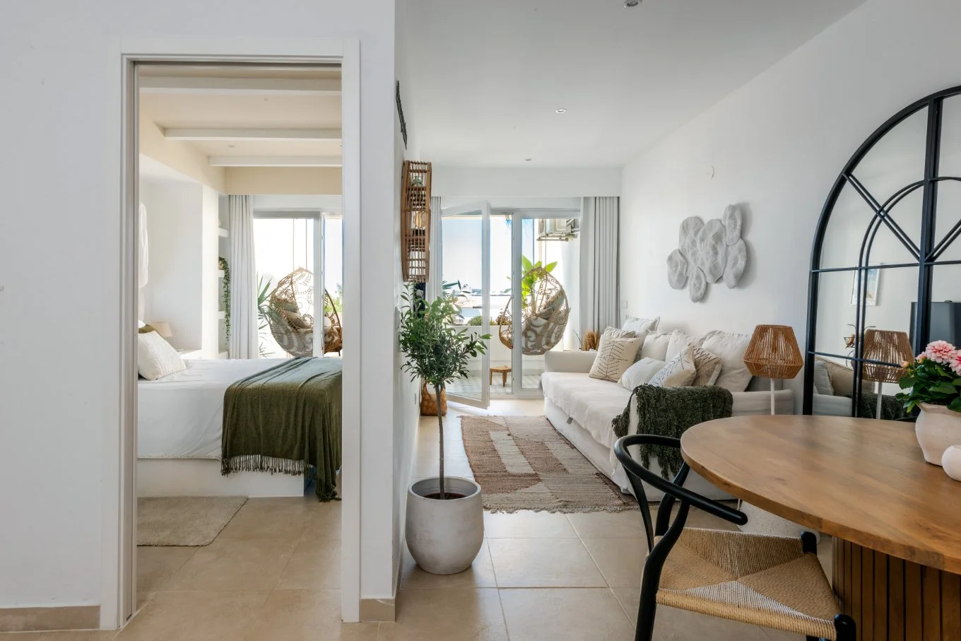 Interior of a modern, bright apartment featuring a bedroom with a green blanket on the bed, and a living area with white sofas, hanging chairs, potted plants, and decorative wall art.