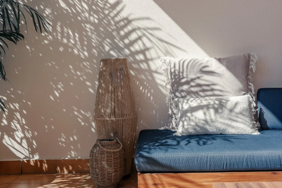 Sunlight casting patterns of shadows from a wicker lamp and a plant onto a white wall, next to a blue-cushioned sofa with two white throw pillows.