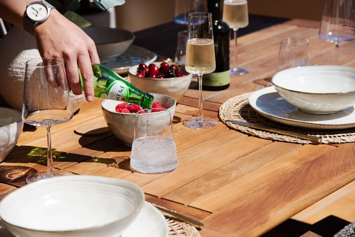 A person is pouring sparkling water into a glass on a wooden dining table set with bowls of fruit, wine bottles, wine glasses, and empty plates.