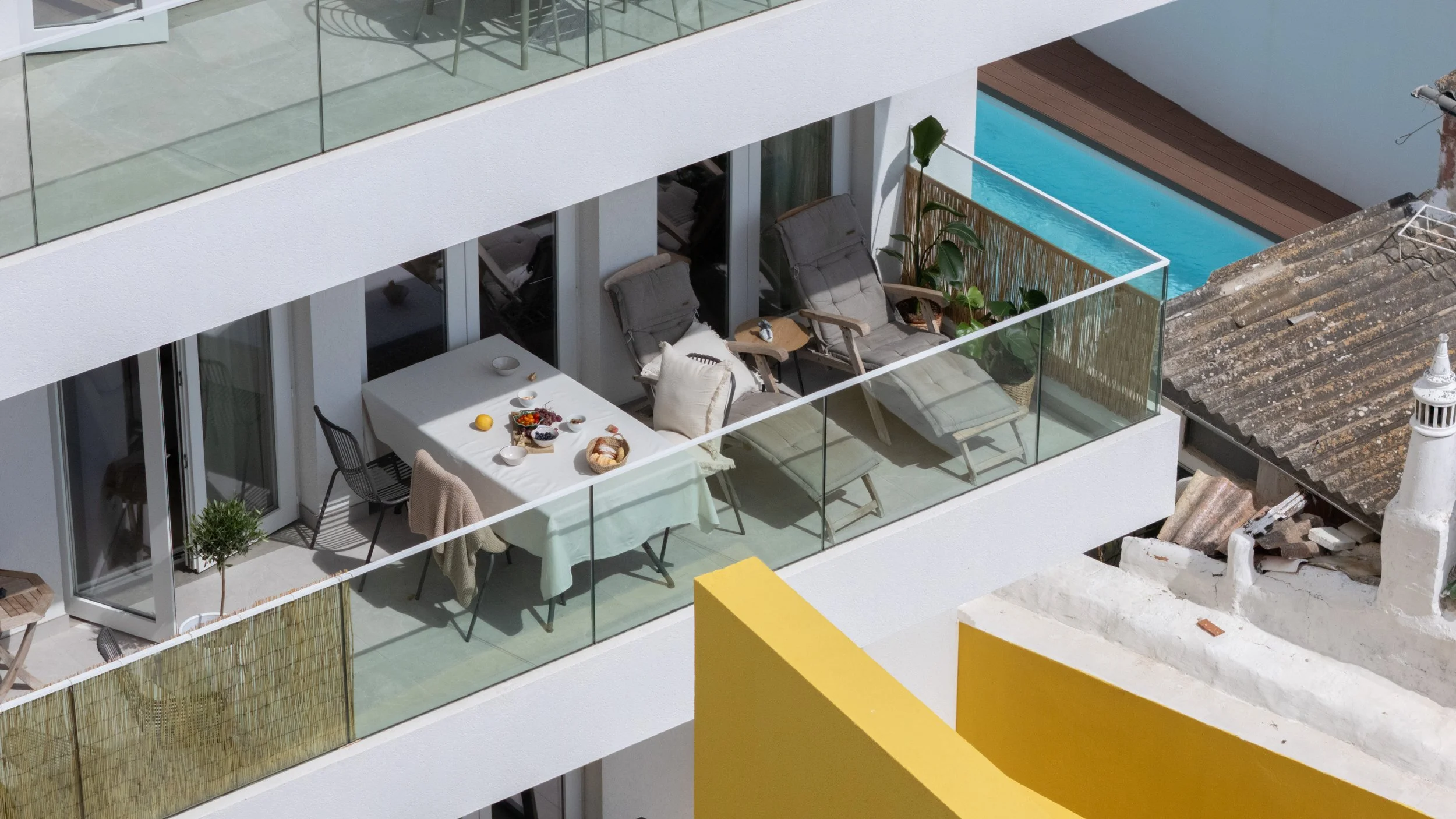Balcony with outdoor furniture, chairs, a table with breakfast items, potted plants, glass railing, and neighboring building with a rooftop and a small pool.