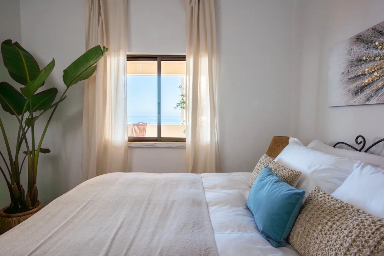 A bedroom with a large bed, decorative pillows, beige bedspread, potted plant, and window with cream curtains overlooking a balcony and ocean. Holiday home in Lagos Algarve