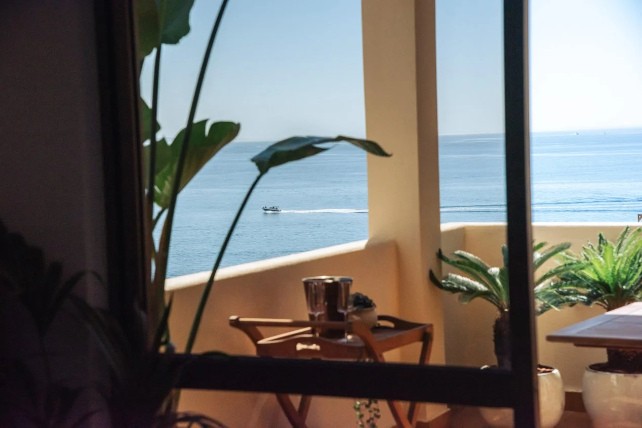 View of the ocean from a balcony, seen through a partially open sliding glass door with indoor plants in the foreground, a small table on the balcony with a plant and metal cups. Holiday home in Lagos Algarve